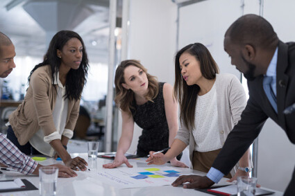 The CEO of a business startup gathers her multiethnic team around the conference table to finalize the launch. She is standing up and smiling as she goes over notes with her team.