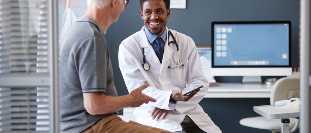 Waist up portrait of smiling African American doctor wearing lab coat and holding tablet while talking to senior patient during consultation in clinic, copy space