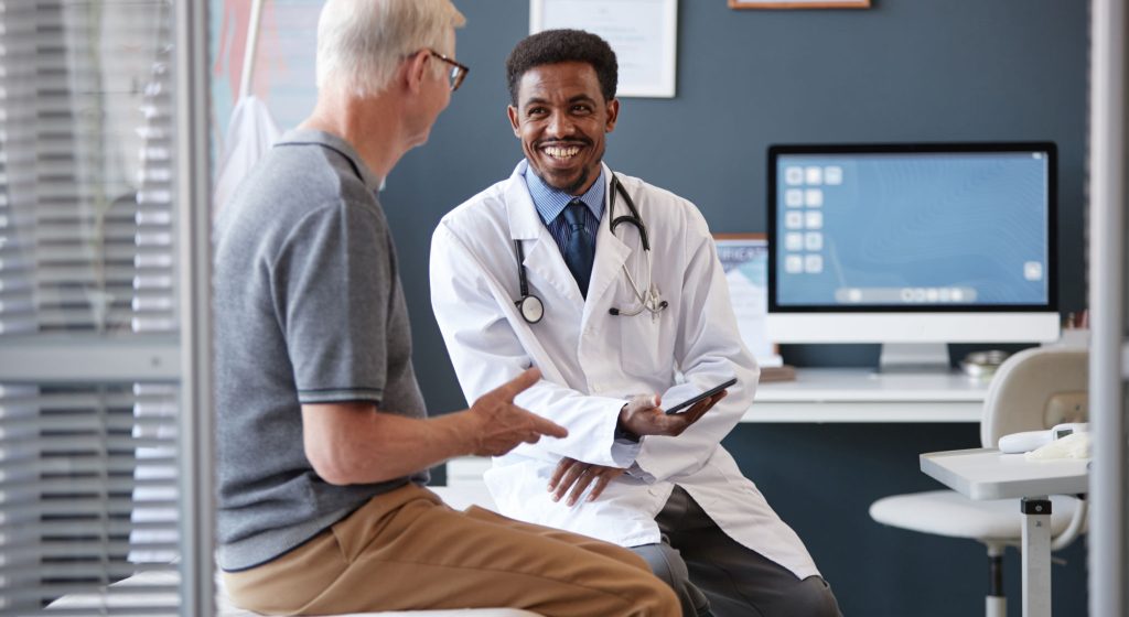 Waist up portrait of smiling African American doctor wearing lab coat and holding tablet while talking to senior patient during consultation in clinic, copy space